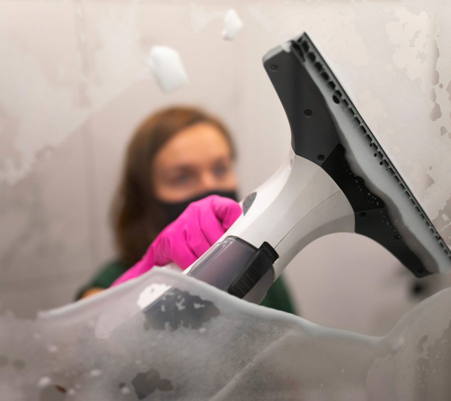 Female washes shower glass with electric glass cleaner. Bathroom cleaning process concept